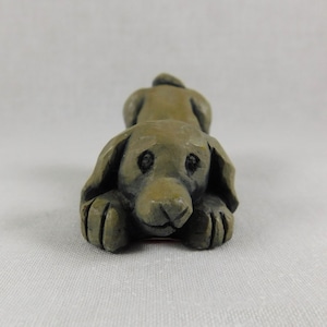 May include: A small, grey, ceramic sculpture of a dog lying down with its head resting on its paws. The dog has a realistic appearance with detailed features, including eyes, nose, and paws.