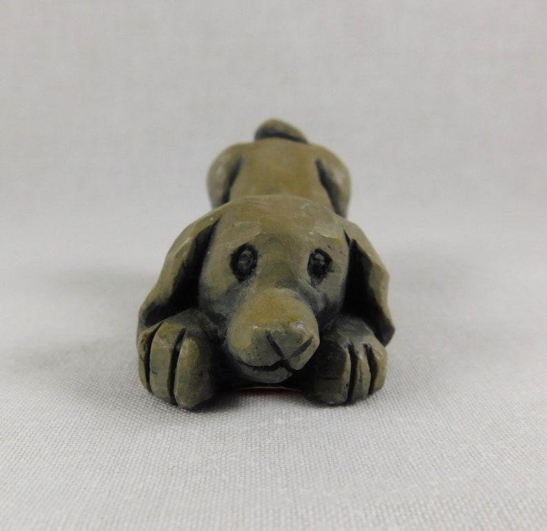 May include: A small, grey, ceramic sculpture of a dog lying down with its head resting on its paws. The dog has a realistic appearance with detailed features, including eyes, nose, and paws.