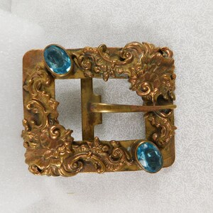 K&ouml;nnte beinhalten: Eine goldfarbene Metallschnalle mit verzierten floralen Designs und zwei blauen Glassteinen.