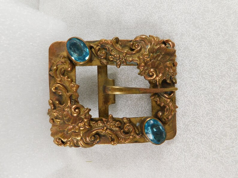 K&ouml;nnte beinhalten: Eine goldfarbene Metallschnalle mit verzierten floralen Designs und zwei blauen Glassteinen.
