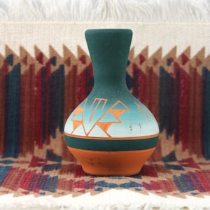 May include: A small, hand-painted ceramic vase with a teal neck and a rounded orange base. The body features a geometric design in orange and white against a light blue gradient. The vase is displayed on a patterned textile.