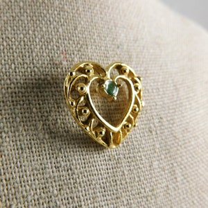 May include: A gold heart-shaped pendant with a green gemstone in the centre. The pendant has a delicate filigree design.