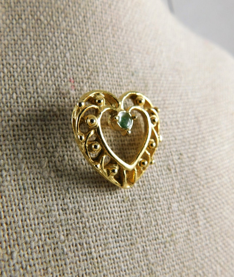 May include: A gold heart-shaped pendant with a green gemstone in the centre. The pendant has a delicate filigree design.