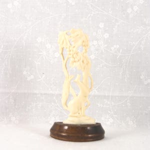 May include: Ivory-colored figurine of a fox reaching up towards a tree with grapes. The fox and tree are carved in detail. The figurine is mounted on a dark brown, oval-shaped wooden base. The background is a white floral pattern.