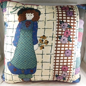 May include: A decorative pillow with a patchwork design featuring a girl in a blue dress holding a bouquet of flowers. The background is a cream color with a brown and white checkered pattern.