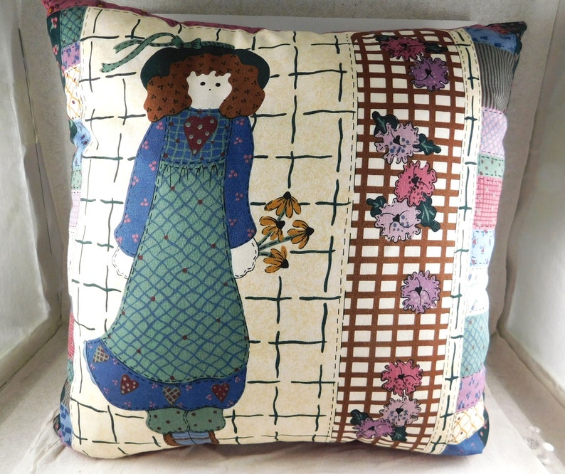 May include: A decorative pillow with a patchwork design featuring a girl in a blue dress holding a bouquet of flowers. The background is a cream color with a brown and white checkered pattern.