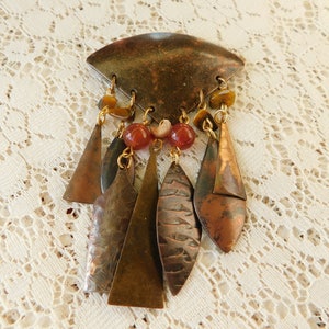 May include: A large, triangular, copper-toned brooch with a dangling chain of copper and gold-toned metal pieces, red and brown beads, and a shell-like bead.