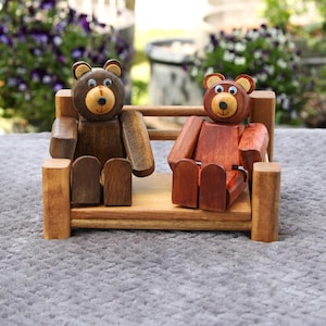 May include: Two wooden bear figurines sitting on a small wooden bench. One bear is dark brown, the other is reddish-brown. Both have tan faces and ears. The bench is light brown and has a simple design. The background is blurred with greenery.