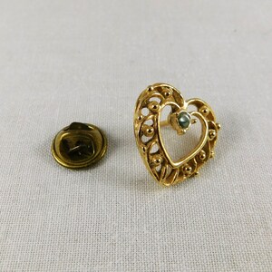 May include: A gold heart-shaped pin with a green gemstone in the centre. The pin has a detailed design with small beads around the edge.