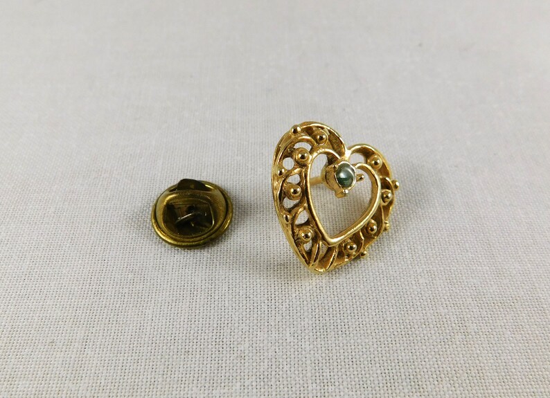 May include: A gold heart-shaped pin with a green gemstone in the centre. The pin has a detailed design with small beads around the edge.