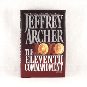 May include: A hardcover book titled "The Eleventh Commandment" by Jeffrey Archer. The cover is primarily dark red with the title in large white letters. The author's name is at the top, and the book features gold-colored accents.