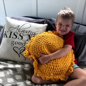 May include: A mustard yellow, chunky knit pillow with a scalloped edge is held by a child. A decorative pillow with the text "Always Kiss Me Goodnight" is visible in the background.