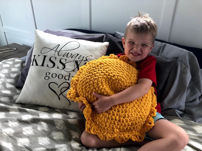 May include: A mustard yellow, chunky knit pillow with a scalloped edge is held by a child. A decorative pillow with the text "Always Kiss Me Goodnight" is visible in the background.