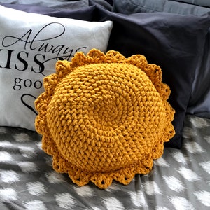 May include: A round, mustard-yellow crochet pillow with a scalloped edge. A white pillow with the text "Always KISS good" in black script is behind it. The pillows are on a bed with gray and white patterned sheets and dark gray pillows.
