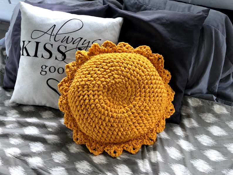 May include: A round, mustard-yellow crochet pillow with a scalloped edge. A white pillow with the text "Always KISS good" in black script is behind it. The pillows are on a bed with gray and white patterned sheets and dark gray pillows.