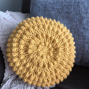 May include: A round, mustard-yellow crochet pillow with a textured, bobble stitch design. The pillow is resting on a gray blanket and a dark brown surface, with a blue pillow in the background. The handmade pillow adds a cozy touch.