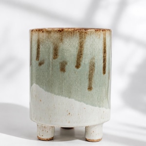 May include: A cylindrical ceramic planter featuring a three-legged base. The upper portion displays a sage green glaze with brown vertical drips, while the lower section is finished in a speckled white glaze that transitions into the base.