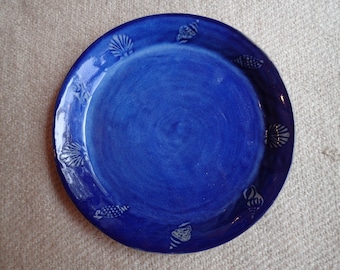 Peut inclure: Une assiette en céramique bleue avec un bord blanc et un motif de coquillages. L'assiette est décorée de coquillages blancs autour du bord.
