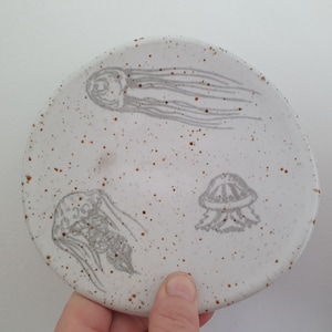 May include: A white ceramic plate with a speckled brown glaze features three hand-drawn jellyfish in grey. The plate is ideal for serving snacks or appetizers.