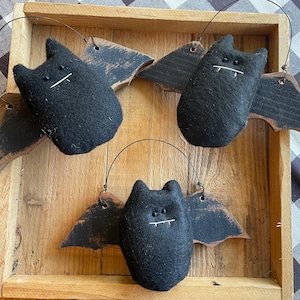 May include: Three black felt bats with wooden wings hanging in a wooden crate. The bats have stitched eyes and mouths.