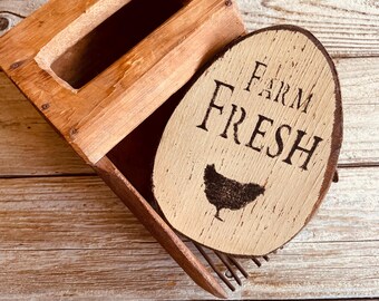 May include: A wooden egg basket and a wooden egg-shaped sign. The sign reads "FARM FRESH" with a silhouette of a chicken. The basket is rectangular with a handle and is made of light brown wood.