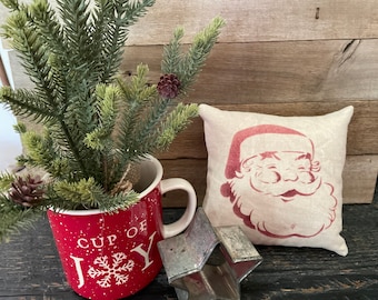 May include: A red mug with white polka dots and the text "Cup of Joy" with a snowflake design. A small pine tree sprig is in the mug. A white pillow with a red Santa Claus face design is on a wooden surface.