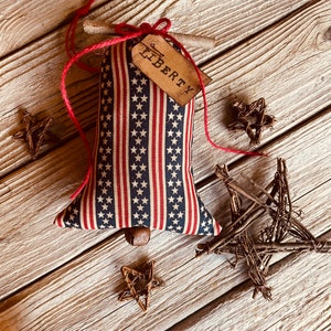 picture shows a red,, white, and blue fabric bell with a brown tag that says 'LIBERTY' hanging from it. The bell is surrounded by four twig stars on a wood background.