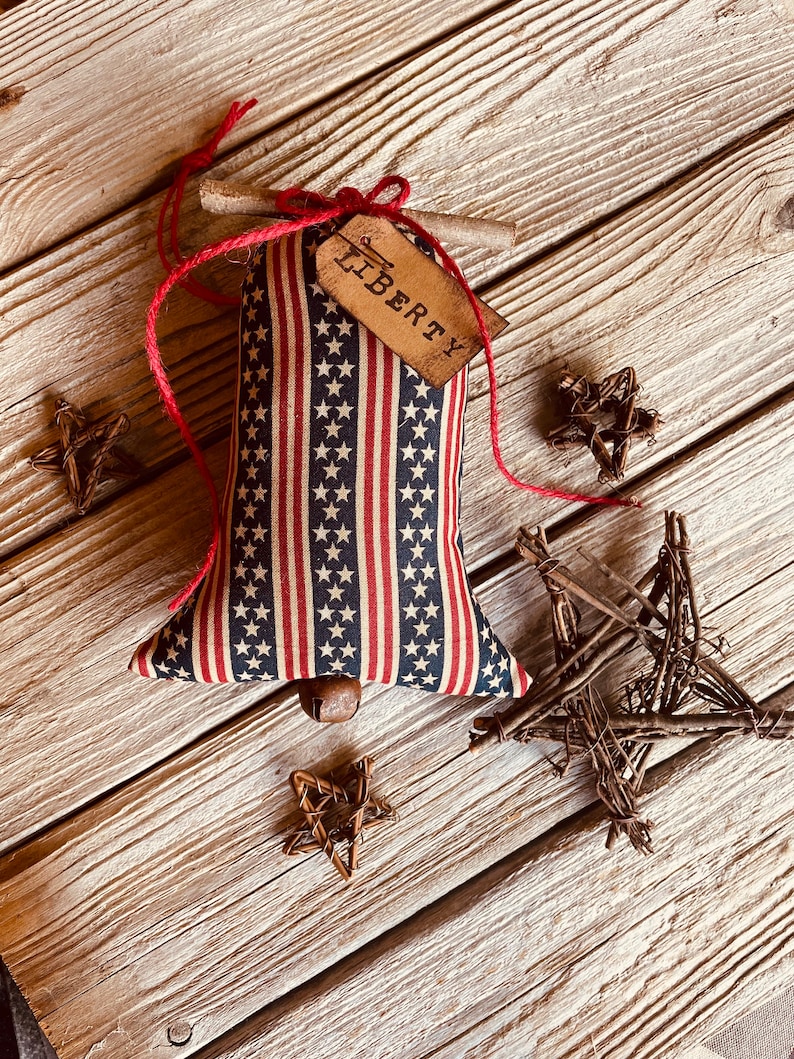 picture shows a red,, white, and blue fabric bell with a brown tag that says 'LIBERTY' hanging from it. The bell is surrounded by four twig stars on a wood background.