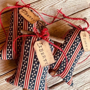 Op de afbeelding: Drie rode, witte en blauwe stoffen bellen met een houten stok en touw. Elke bel heeft een bruin label met het woord "LIBERTY" erop gedrukt.