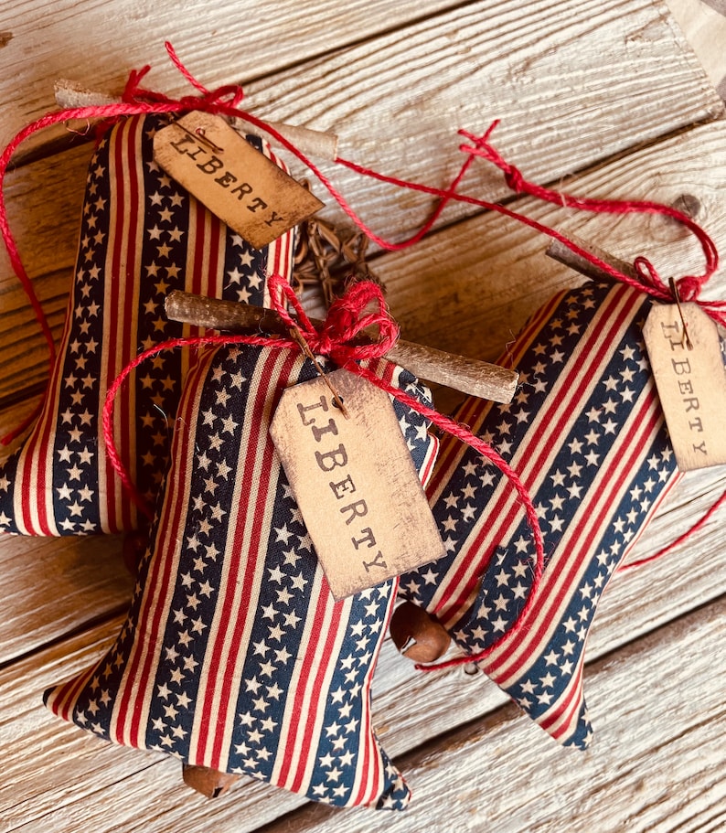 Op de afbeelding: Drie rode, witte en blauwe stoffen bellen met een houten stok en touw. Elke bel heeft een bruin label met het woord "LIBERTY" erop gedrukt.