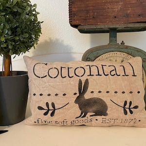 以下が含まれることがあります： 黒い文字で「Cottontail」と書かれたベージュの枕。枕には、ウサギの黒いシルエットと「fine soft goods. EST. 1872.」という文字も書かれている。