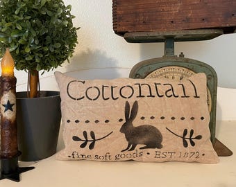 Może przedstawiać: Beżowa poduszka z napisem "Cottontail" czarnymi literami. Poduszka zawiera również czarną sylwetkę królika i napis "fine soft goods. EST. 1872."