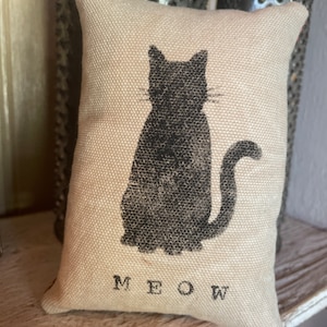 May include: A beige pillow with a black silhouette of a cat and the word "MEOW" printed on the bottom.