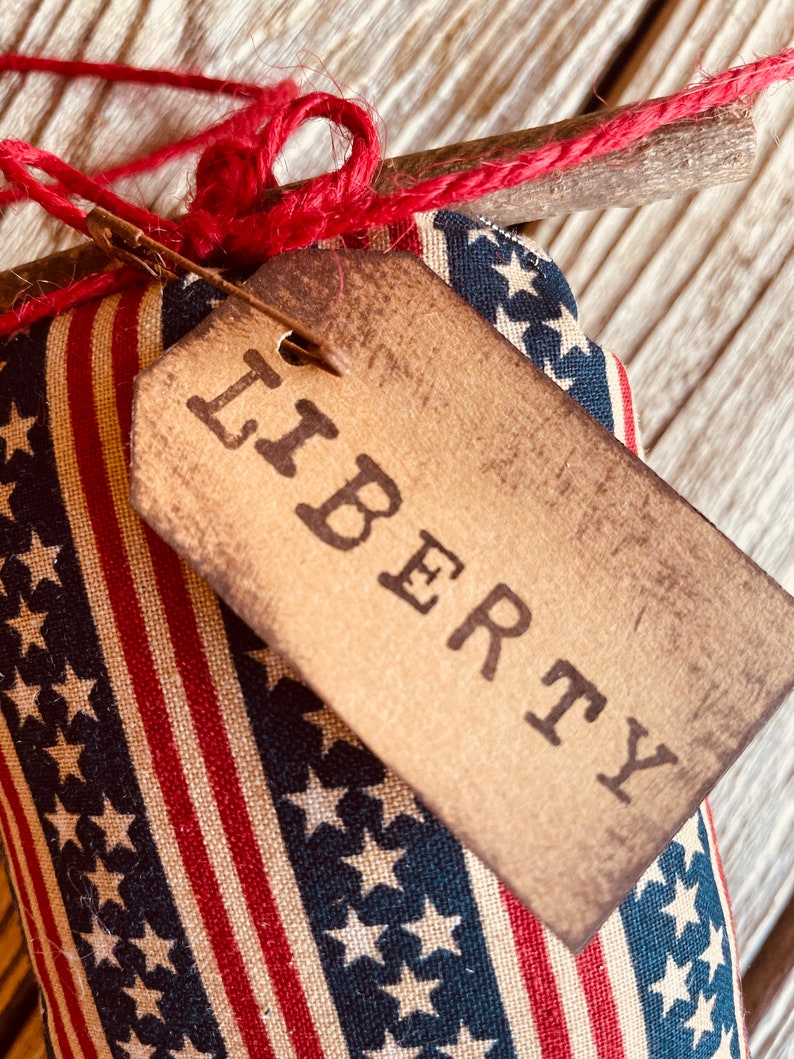 Op de afbeelding: Een klein, handgemaakt kussen met een rood, wit en blauw stoffen ontwerp met sterren en strepen. Het kussen heeft een bruin label met de tekst "LIBERTY".