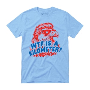 May include: Light blue t-shirt featuring a red eagle wearing sunglasses with a star and the text "WTF IS A KILOMETER?" in blue. The eagle graphic has a retro design. The shirt is made of a soft material.