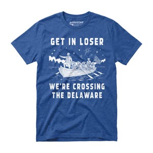 May include: Blue t-shirt with white graphic text and illustration. The text reads "GET IN LOSER" above a depiction of a boat crossing a river, with the text "WE'RE CROSSING THE DELAWARE" below. The illustration includes a flag and fireworks.
