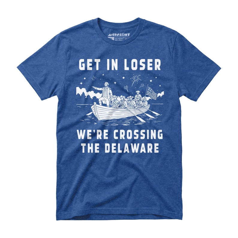 May include: Blue t-shirt with white graphic text and illustration. The text reads "GET IN LOSER" above a depiction of a boat crossing a river, with the text "WE'RE CROSSING THE DELAWARE" below. The illustration includes a flag and fireworks.