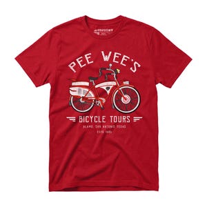 May include: Red t-shirt with a vintage bicycle graphic. The design features a red and white bicycle with the text "PEE WEE'S BICYCLE TOURS" above and "ALAMO. SAN ANTONIO TEXAS ESTO 1985" below. The shirt is made of a soft material.