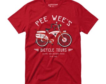 Puede incluir: Camiseta roja con un gráfico de bicicleta vintage. El diseño presenta una bicicleta roja y blanca con el texto "PEE WEE'S BICYCLE TOURS" arriba y "ALAMO. SAN ANTONIO TEXAS ESTO 1985" abajo. La camiseta está hecha de un material suave.