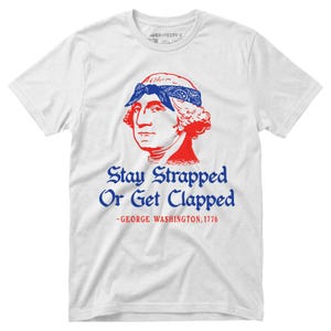 May include: A white short-sleeve t-shirt featuring a red graphic of George Washington wearing a blue bandana. Below the graphic, the blue text reads Stay Strapped Or Get Clapped, followed by the red text ~GEORGE WASHINGTON, 1776.