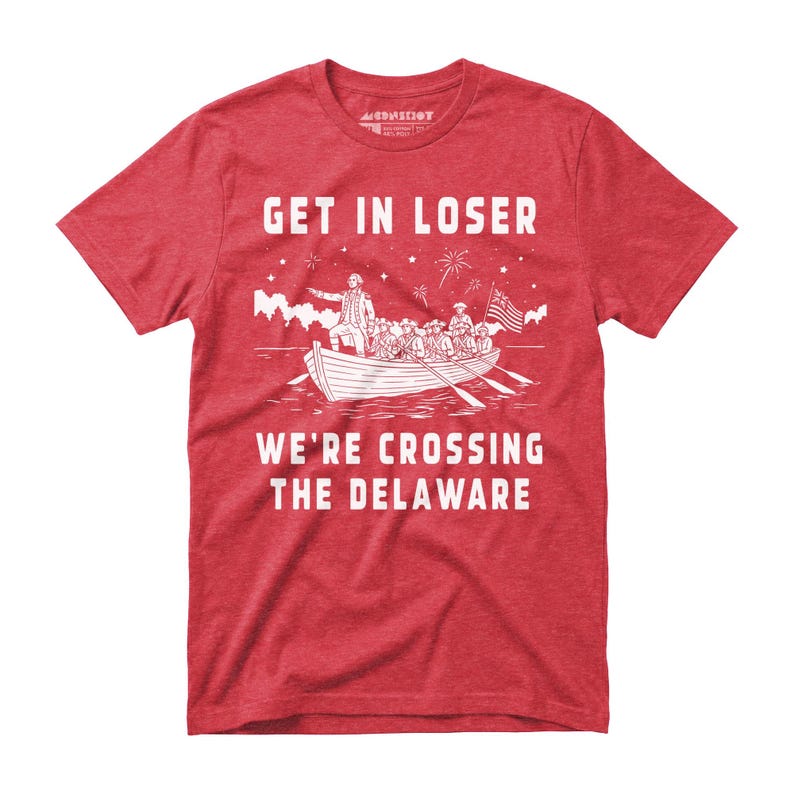 May include: Red t-shirt with a white graphic of a boat crossing a river, featuring a historical scene with the text "GET IN LOSER WE'RE CROSSING THE DELAWARE". The shirt has a crew neck and short sleeves.