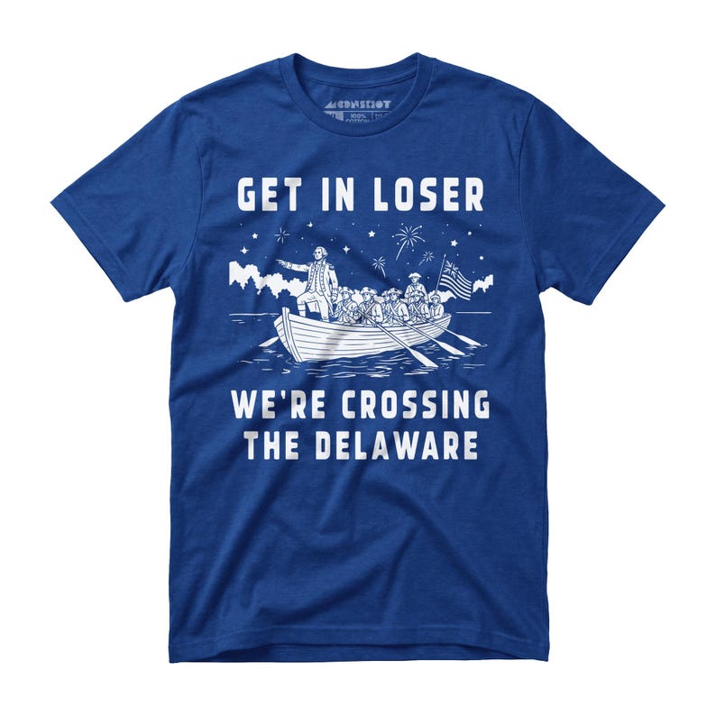 May include: Blue t-shirt with white graphic of a boat crossing water, with text "GET IN LOSER" above and "WE'RE CROSSING THE DELAWARE" below. The boat has people and an American flag.