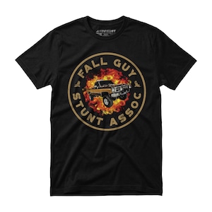 May include: Black t-shirt featuring a circular graphic with a vintage truck engulfed in flames. The graphic includes the text "FALL GUY STUNT ASSOC" in a circular design. The shirt has short sleeves and a crew neck.