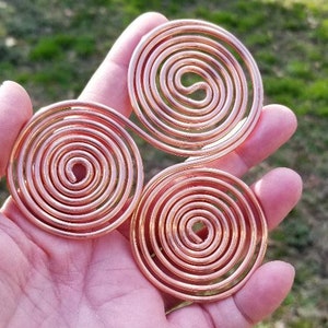 Pode incluir: Três ornamentos em espiral de arame de cobre, cada um com uma abertura central. As espirais são seguradas em uma mão contra um fundo verde desfocado. O cobre tem um tom marrom-avermelhado quente.