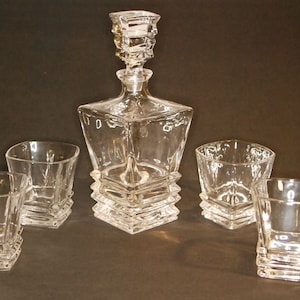 May include: A set of five clear glass decanter and glasses with a geometric design. The decanter has a square base and a tall, narrow neck. The glasses are short and wide, with a square base.