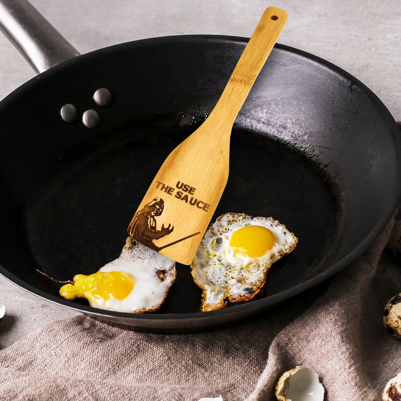 A wooden spatula is lying in the pan. Bamboo spatula is engraved with Darth Vader and the inscription - Use the sauce. The engraved spatula is designed for Star Wars fans.