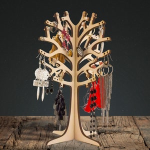 Puede incluir: Soporte de árbol de madera para pendientes con múltiples ramas y agujeros para colgar pendientes. El soporte está decorado con varios pendientes en plata, oro, negro y rojo.