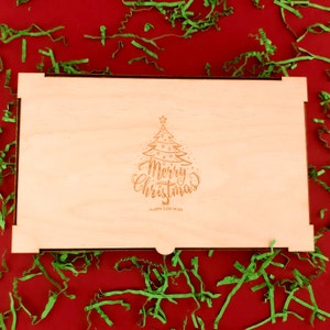 The wooden engraved box is placed on a red background with green small decorations. Merry Christmas with a Christmas tree is engraved on the box.