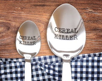 May include: Two silver spoons with the text "Cereal Killer" engraved on them. The spoons are on a wooden surface with a blue and white checkered cloth.