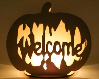 May include: A wooden pumpkin-shaped candle holder with a cutout design that spells out "Welcome". The candle holder is lit and glowing.